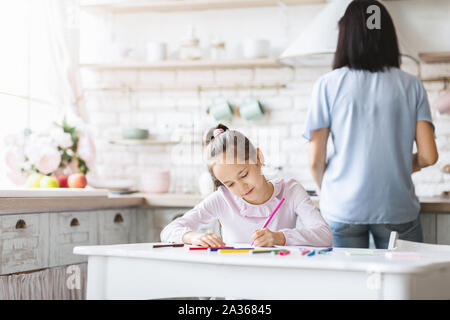 La scolaretta facendo i compiti in cucina mentre la madre di cottura degli alimenti Foto Stock