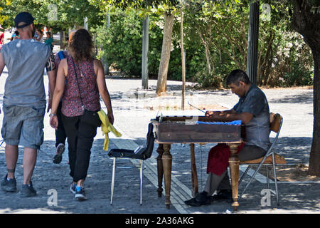 Atene, Grecia, giugno 04.2016.i turisti ad Atene a piedi passato un musicista di strada cercando di farli divertire da seduti all'ombra di un ulivo. Foto Stock