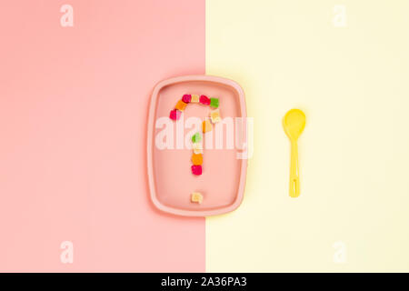 Minimalista di corteccia di piatto con una rosa di plastica sulla piastra che è un punto interrogativo di canditi di ananas e un cucchiaio di giallo in un giallo-rosa backgro isolato Foto Stock