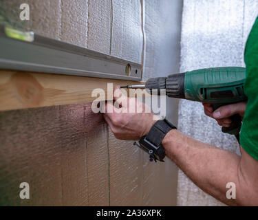 La mano di sesso maschile con l aiuto di un trapano elettrico-cacciavite e una vite autofilettante fissa la barra di legno sul quale giace il livello alla parete del th Foto Stock