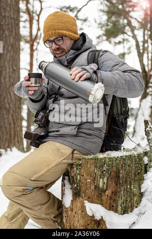 Un bel uomo barbuto con gli occhiali con una macchina fotografica e uno zaino seduto su un moncone nella foresta versa il tè caldo da un thermos in una tazza e sorriso Foto Stock