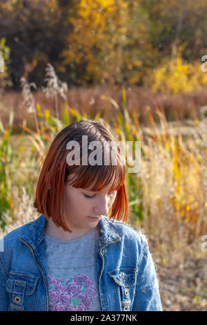 Una ragazza sta con la sua testa piegata in una giornata di sole contro lo sfondo di un bosco d'autunno. La ragazza ha i capelli rossi. Legno e canne in background Foto Stock