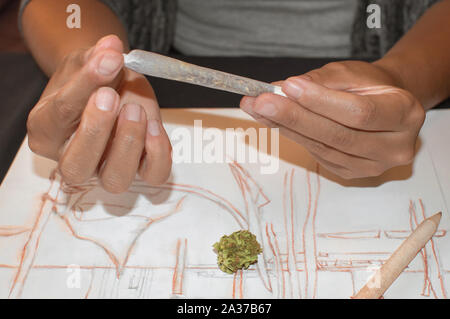 Giovane donna laminazione di un giunto di marijuana durante una pausa dopo il disegno. Concetto di droga nell'arte. Sfondo con la cannabis bud, matite e schizzi Foto Stock