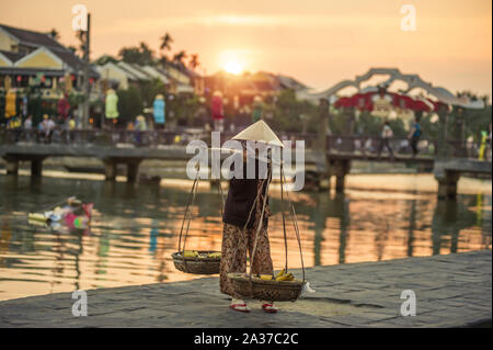 Hoi An, Quang Nam Provincia, Vietnam - Febbraio 24, 2011: venditore ambulante che porta frutti di cestelli in banche di Thu Bon River alla antica città di Hoi An Foto Stock