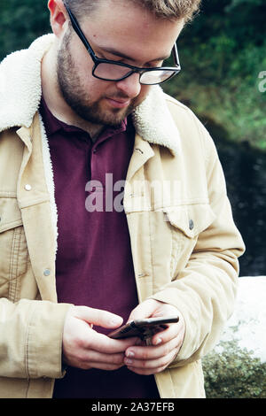 Un primo piano di un elegante e bello giovane maschio con la barba e i bicchieri controllando il suo telefono cellulare per i messaggi e i social media all'aperto Foto Stock