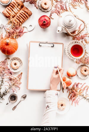 Donna femmina scrive a mano su carta bianca pezzo di carta sulla scheda di clipping sull accogliente casa autunno scrivania con impostazione di tè, dolci, autunno fiori e foglie, Foto Stock