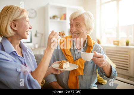 Ritratto di felice coppia senior di mangiare la torta insieme e ingannare intorno godendo di fine settimana a casa, scena illuminata da luce solare Foto Stock