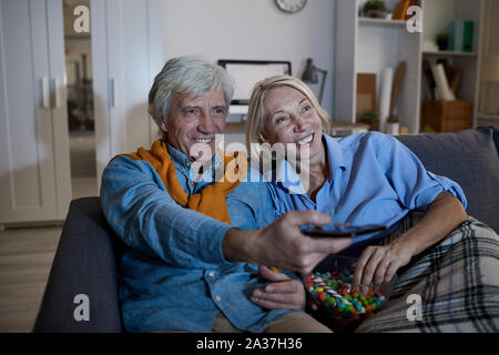Ritratto di sorridente coppia senior guardando la TV insieme seduti sul comodo divano nel buio, spazio di copia Foto Stock