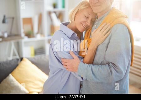 Ritratto di tagliate di amorevole coppia senior abbracciando teneramente mentre in piedi nel soleggiato soggiorno a casa, spazio di copia Foto Stock