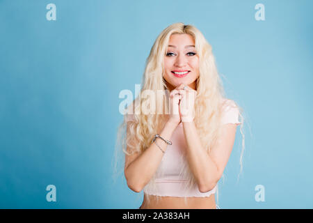 Felice giovane donna bionda in amore, mantenendo le mani sulle guance, gioiosamente sorridente con denti bianchi, guardando in alto, vestito di rosa pallido tee shirt, isolate su Foto Stock