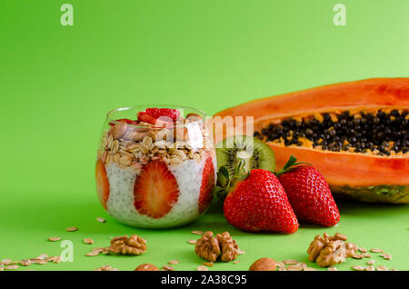 Detox e sana colazione superfoods concetto. Chia budino di yogurt con fragole, muesli e frutta secca in un bicchiere e papaia con kiwi su verde Foto Stock