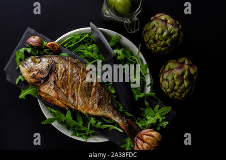 Vista superiore del cotto Orate di mare su una ciotola con rucola, carciofi e coltello su sfondo nero. Mediterranean Food concept. Foto Stock