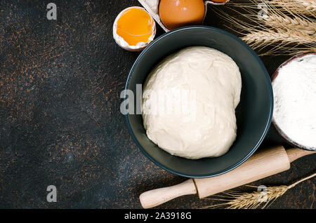 Materie la pasta lievitata in una terrina e ingredienti, uova, farina, spighe di grano scuro sul tavolo di cucina e panetteria concetto, Foto Stock