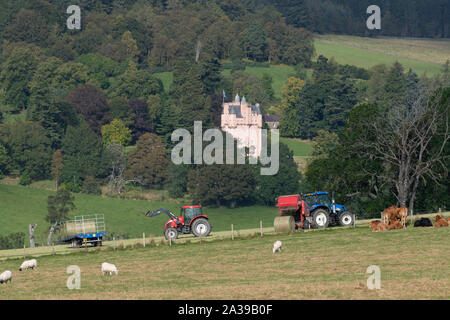 Un trattore e l'imballatrice con il deposito di una nuova balla di fieno come un secondo trattore carichi di balle su una balla di attesa rimorchio nella parte anteriore del Castello di Craigievar in Aberdeenshire Foto Stock