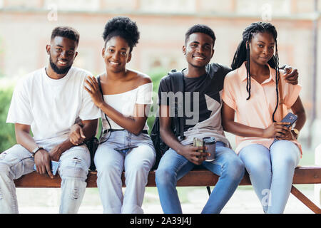 Un gruppo di giovani amici africani appendere fuori insieme all'aperto su un banco e ridere mentre discutono Foto Stock