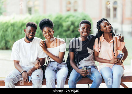 Un gruppo di giovani amici africani appendere fuori insieme all'aperto su un banco e ridere mentre discutono Foto Stock