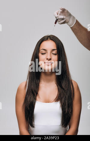 Cosmetologo in lattice Guanti di cuoio capelluto facendo la mesoterapia a brunette bella donna con capelli lunghi isolati su grigio Foto Stock