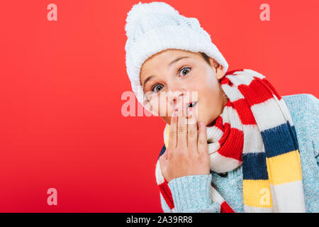 Sorpreso capretto nel cappello e sciarpa che copre la bocca con la mano isolato su rosso Foto Stock