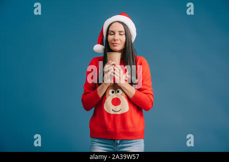 Felice ragazza con gli occhi chiusi in santa hat e Natale maglione azienda Caffè di andare, isolato su blu Foto Stock