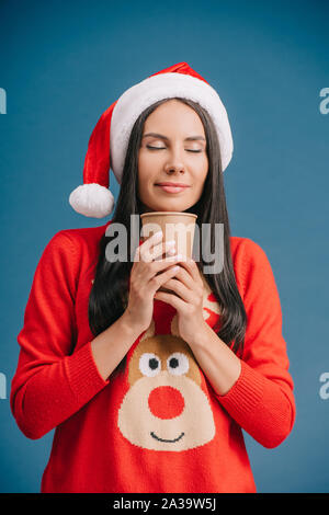 Donna attraente con gli occhi chiusi in santa hat e Natale maglione azienda Caffè di andare, isolato su blu Foto Stock