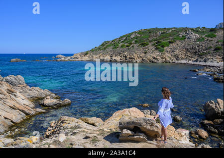 La donna vista posteriore in un vestito blu si appoggia su una roccia e si affaccia al mare di colori in un giorno chiaro Foto Stock