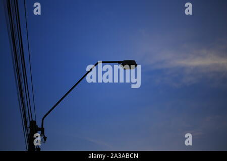 Lampada post silhouette. via la luce su un palo in cemento di linee di alimentazione. Delineare delle infrastrutture contro twilight ora oscurato il cielo blu e sparse cloud. Foto Stock