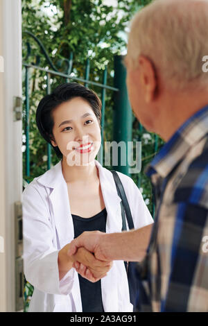 Piuttosto sorridente giovane vietnamita medico agitando la mano di uomo senior dopo la visita di lui a casa e discutere il processo di terapia Foto Stock