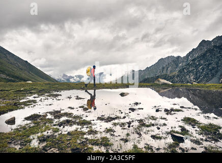 Turistico con zaino giallo è a piedi vicino al lago con la sua riflessione a valle verde a Karakol national park, il Kirghizistan Foto Stock