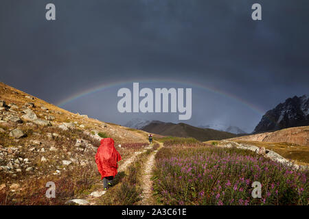 Turista nella Red Rain cover è a piedi giù per la strada di montagna di arcobaleno a sfondo Foto Stock