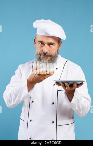 Ritratto di barbuto senior chef tenendo la piastra a vuoto mentre in piedi contro lo sfondo blu e guardando la telecamera, mock up modello Foto Stock