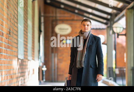 Giovane uomo in soprabito e sciarpa avvicinando la fotocamera tirando una valigia dietro di lui lungo un passaggio esterno Foto Stock