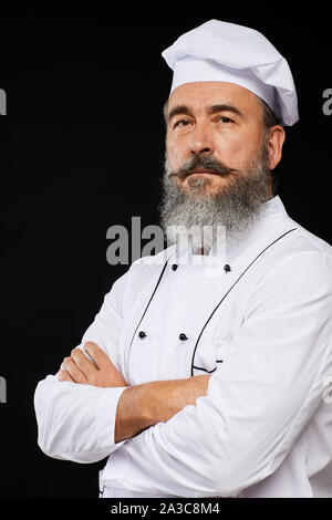 Vita professionale su ritratto del barbuto senior chef che posano su sfondo nero in piedi con le braccia incrociate Foto Stock
