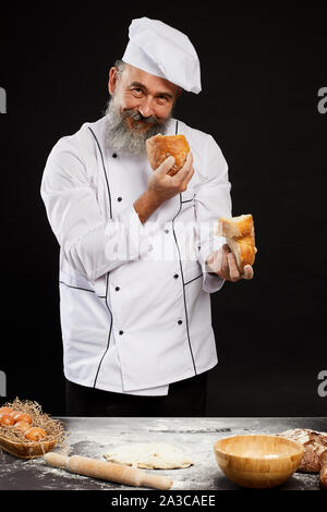 Ritratto di Allegro barbuto baker holding fresh pagnotta di pane guardando la fotocamera mentre si posa su sfondo nero Foto Stock