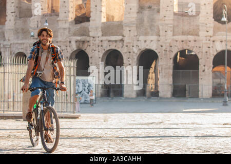 Felice giovane turista in Bicicletta Equitazione indossando maglietta e hat al Colosseo a Roma, Italia di sunrise. Foto Stock