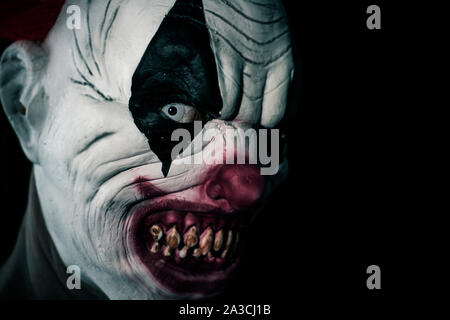 Primo piano di una paura male clown con il bianco degli occhi, denti sanguinante e un aspetto minaccioso fissando l'osservatore, contro uno sfondo nero Foto Stock