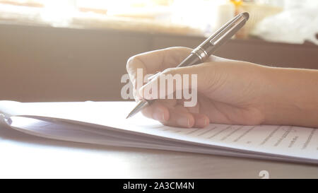 Close up donna mani di firma o la scrittura di un documento su un foglio di carta bianca con la penna nera al mattino Foto Stock