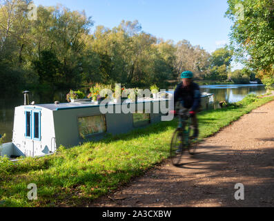 Uomo in bicicletta lungo il Tamigi percorso in corrispondenza di velocità, passando una barca stretta in un pomeriggio autunnale accanto al Fiume Tamigi a Sonning-Berkshire, England, Regno Unito Foto Stock