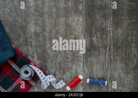 Accessori per il cucito, jeans e plaid tessuto su di un legno scuro dello sfondo. Tessuto, fili di cucitura, un ago per cucire e per centimetro. top view, flatlay Foto Stock