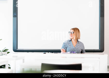 Giovani pensieroso attraente professore con gli occhiali in abiti formali di insegnamento in classe e a sostenere l'esame. seduta vicino a Lavagna. Foto Stock