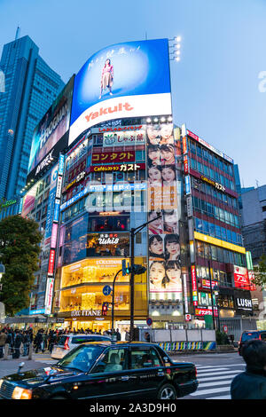 Tokyo, Shibuya. Taxi passando al di sopra di attraversamento pedonale con L'Occitane Cafe, Ushi 8 e vari altri in background torre angolare blocco. Foto Stock