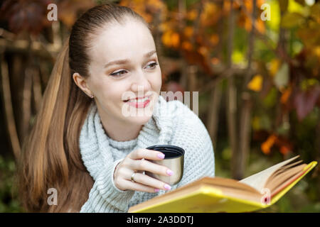 Una ragazza legge un libro e bevande di tè da un thermos. Sorriso. Autunno nel parco. Stile di vita concetto, autunno, relax e lettura. Foto Stock