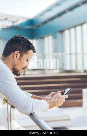 Giovane uomo appoggiato sulla ringhiera mentre si utilizza il telefono cellulare Foto Stock