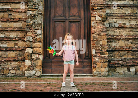 Adorabile bambino con zaino sventolando giocattolo con il mulino a vento di carta permanente sulla strada di età contro il muro di pietra con grande porta di legno in Toscana Italia Foto Stock