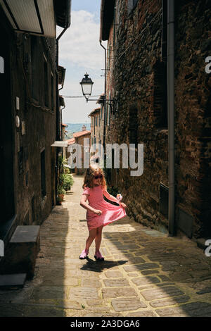 Allegro ragazza in abito rosa e occhiali da sole di girare intorno mentre si cammina lungo la strada pedonale con case in mattoni Foto Stock