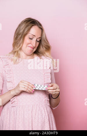 Giovane donna incinta indossando abiti rosa guardando il blister della medicina compresse in confusione medita di prendere pillole durante la gravidanza o non. Studio Foto Stock