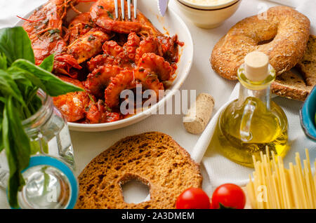 Close-up di gamberi cotti sgusciati e il polpo in una ciotola bianco. Pasta, friselli, olio di oliva, basilico e pomodori sul tavolo. Mediterranean Food concept. Foto Stock