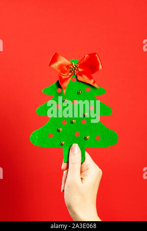 Feltro albero di Natale fatto da mani con tallone d'oro dei giocattoli e un fiocco rosso nella parte superiore in una mano dura su uno sfondo di colore rosso. Artigianato di natale Foto Stock