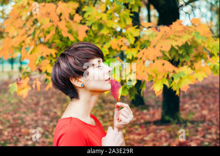 Autunno Romantico bellezza. Ritratto di donna carina in abito rosso con foglie di autunno nel parco all'aperto nella giornata di sole. Stagionale umore sognante. Spazio di copia Foto Stock