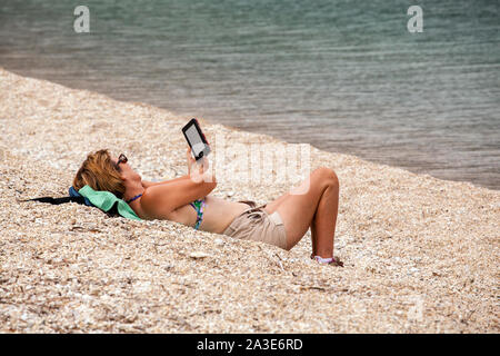 Donna sdraiata su una spiaggia di ciottoli la lettura di un libro di kindle reader Foto Stock