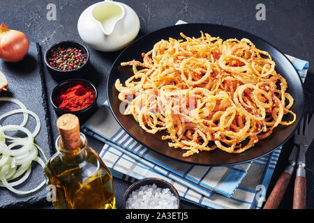 Il francese croccante di anelli di cipolla fritti e stringhe su una piastra nera su una tabella di calcestruzzo con ingredienti, orizzontale vista da sopra Foto Stock